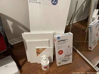 Photo showing large wooden easel behind stacked blank canvases, two paint kits in original packaging, and a white pump bottle, positioned on floor by mirror and chair.