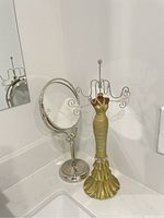 Two images showing a silver-tone double-sided pedestal countertop mirror and a gold-tone ornamental jewelry stand shaped like a dress with multiple arms for holding jewelry.