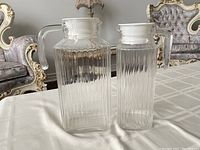 Two clear glass jugs with white lids on table. Both jugs have vertical ridges. One jug has a slightly damaged lid.