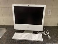 Front view of Apple iMac with wired keyboard and power cable showing intact unit with cracked screen bottom left corner.
