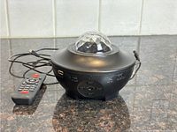 Front and top view of black round light projector with crystal-like dome, USB power cable, and remote control on countertop