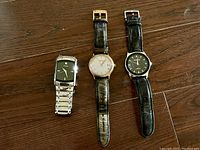 Front view of three men's watches laid out on wooden surface: a silver-tone Fossil bracelet watch, a gold-tone Fossil leather strap watch with Roman numerals, and a Montine black leather strap watch.