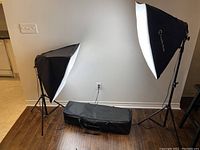 Two set up soft box lights with lit bulbs on adjustable tripods and carrying bag on floor