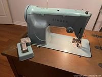 Light blue standalone Singer sewing machine with foot pedal on wooden floor.