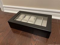 Closed black watch box with glass lid on wooden floor, showing compartments through glass
