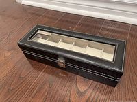Closed black watch box with glass top and white stitching displayed on hardwood floor.