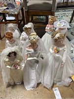 Seven Ashton Drake bridal dolls in white and cream gowns, posed standing on floor with chairs behind.