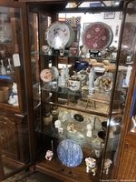 Wide view of display cabinet with various Asian style porcelain plates, figurines, and decorative items arranged on three glass shelves.