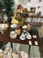 Photo of all lot contents arranged on table including canisters, tea cups on saucers, faux flowers, salt lamp, plant stand, and egg tray.