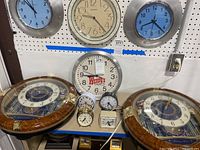 Photo showing a rack with 3 wall clocks mounted above a table with 6 tabletop and alarm clocks arranged, including two large Seiko musical wall clocks, a Wendy's metal rim clock, and smaller alarm clocks on table.