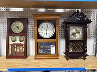 Shelf display of 3 wall clocks: Daniel Dakota clock with winter scene, restaurant facade clock, and dark wood ornate clock.