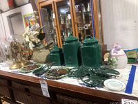 Overview of lot on a white lace tablecloth including anniversary clocks under glass domes, green ceramic canisters, dark green cast iron trivets, and a white ceramic jar with bird lid.
