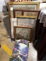 Photo showing all nine framed prints stacked against a wall, displaying variety in sizes, frames, and art styles including needlepoint style and nature themes
