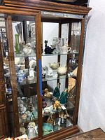 Full view of wooden display cabinet with glass shelves showing various porcelain, ceramics, and collectibles including teapots, vases, and figurines.