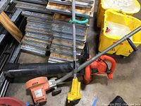 Three electric garden tools: Black & Decker Leaf Hog blower, orange Black & Decker edger, yellow electric weed eater. All placed on floor with other materials around.