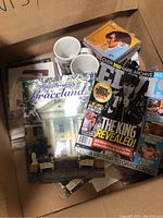 Box containing two white Elvis Presley mugs, magazines including 'Elvis The King Revealed', and a Graceland official guidebook