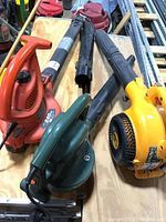 Three leaf blowers on a wooden surface: orange-black Black and Decker LH4500, yellow Poulan Pro BVM200VS, and green-black Black and Decker BV500 with extra accessory.