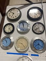 Eight wall clocks arranged on a pegboard, various sizes and frame colors including black, grey, silver, and clear. Faces show a mix of white, blue, and cream with different numeral styles.