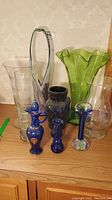 Front view of group of eight glass items on wooden surface including green large vase, clear vases, cobalt blue decanter and two cobalt art glass pieces, black etched vase.