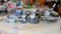 Wide view of table showing blue and white porcelain teacups, saucers, rice bowls, plates, and pinkish lusterware pieces placed on a lace tablecloth.