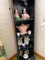 Overview of black shelf unit showing various figurines, china teapot, teacup with saucer, Southern Bell figural bookends, and other decorative items.
