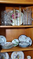 Photo of two crystal pitchers and floral china plates on top shelf, gilded champagne flutes in front