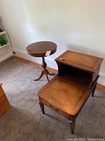 Two tables side by side on carpeted floor near wall; one is a tiered mahogany side table with drawer, the other a round mahogany drum table with tripod brass-capped legs.