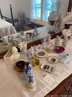 Photo showing collection of assorted figurines, mini bud vases, trinket dishes and other souvenir ceramics arranged on a white covering in front of a mirror.
