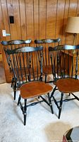 Set of four Hitchcock style chairs with black painted spindle backs and brown wooden seats arranged on carpet against wood-paneled wall