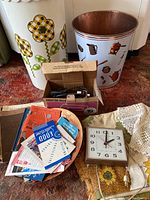 Photo showing the assortment of vintage cookbooks, electric clock, metal bowl, and vintage iron in original box placed on a textured carpet next to flower-themed enamel waste cans