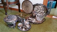 Photo showing silverplated serving pieces including chafing pot, silver trays, condiment sets, flatware and crystal bowl arranged on floor
