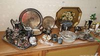 Wide shot of silverplated serving items including teapots, trays, bowls, utensils, vases, and coasters arranged on wooden surface.
