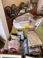 Wide view of assortment of tablecloths, runners, napkins, and small pillows displayed on a couch and shelving area.