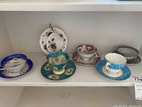 Overview of six intricate teacup and saucer pairs arranged on shelf, various floral and gold details.