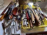 Overview photo showing a wide array of kitchen utensils including carving knives with wooden handles, spatulas, metal potato masher, scissors, and various metal and wooden kitchen gadgets arranged in yellow trays and on a counter.