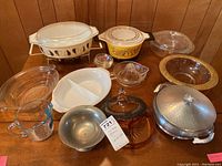 Full lot on wood table showing vintage Pyrex casseroles, serving racks, Anchor Hocking measuring cup, covered candy dish, glass citrus juicer, glass bowls, metal chafing dish.