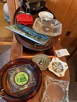 Various vintage ashtrays and trays arranged on furniture showing clear and amber glass ashtrays, commemorative ashtrays with crests, and decorative trays.