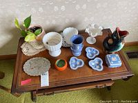 Full view of all items arranged on wooden table including the plant holder, milk glass compotes, Wedgwood Jasperware pieces, candy dish, metal trivets, and fish-shaped vessel.