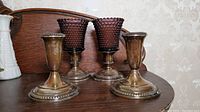 Four candle holders on dark wood surface with carved wall panel behind. Two are sterling silver weighted holders showing tarnish and beaded edge base, two are weighted holders with purple hobnail glass tops.