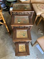 Four wooden nesting tables stacked showing the shadow box glass tops with Asian style floral artwork under the glass and carved wood aprons.