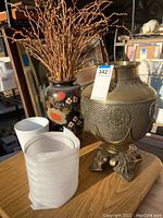 Photo showing the large brass vase with engraved details, the tall black ceramic floral vase with branches, the white ceramic vase, and the clear glass vase on table.