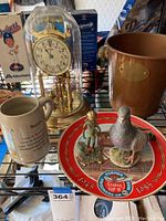 Items on a wire shelf including a Kundo anniversary clock under glass dome, brass figurine, ceramic pigeon on commemorative plate, Bavaria prisoner camp mug, and copper bucket