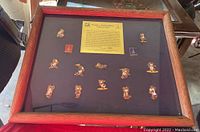 Framed set showing multiple small enamel pins featuring Misha, the mascot of the 1980 Moscow Olympics, mounted on dark backing with wooden frame.
