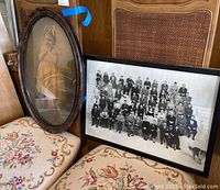 Two framed photos pictured on an embroidered chair; an oval framed vintage photo of a little girl and a large black and white group photo in a rectangular frame.