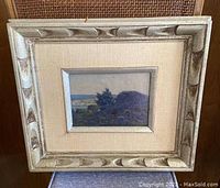 Framed landscape oil painting shown from front. Frame is textured wood with silver finish and beige fabric mat surrounding the painting. Wall background visible.