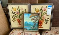 Three framed paintings: two floral paintings on glass and one small nature oil painting, arranged side by side on fabric surface.