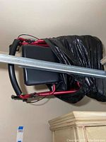 Folded seated walker with red metal frame, black padded seat and backrest, wrapped partially in black plastic bag and mounted high on wall.