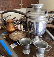 Photo of the entire lot showing the coffee urn in the center, copper coffee pot to the side, pewter cream and sugar containers in front, decorative tray underneath, and small bowl nearby.