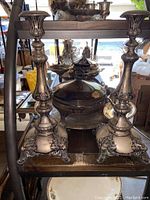 Photo showing two large ornate silver plated candlesticks with floral scroll patterns and a wide square base.