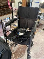 Side and front angle view of black foldable wheelchair showing padded armrests and fabric seating, with black plastic parts and straps on seat.